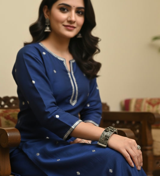 Woman in a blue traditional outfit and wearing bracelet sitting on a wooden chair.