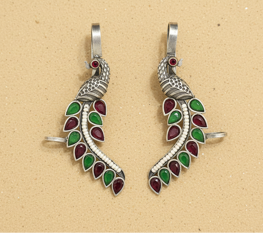 Two silver peacock-shaped pendants with green and red accents on a beige background.