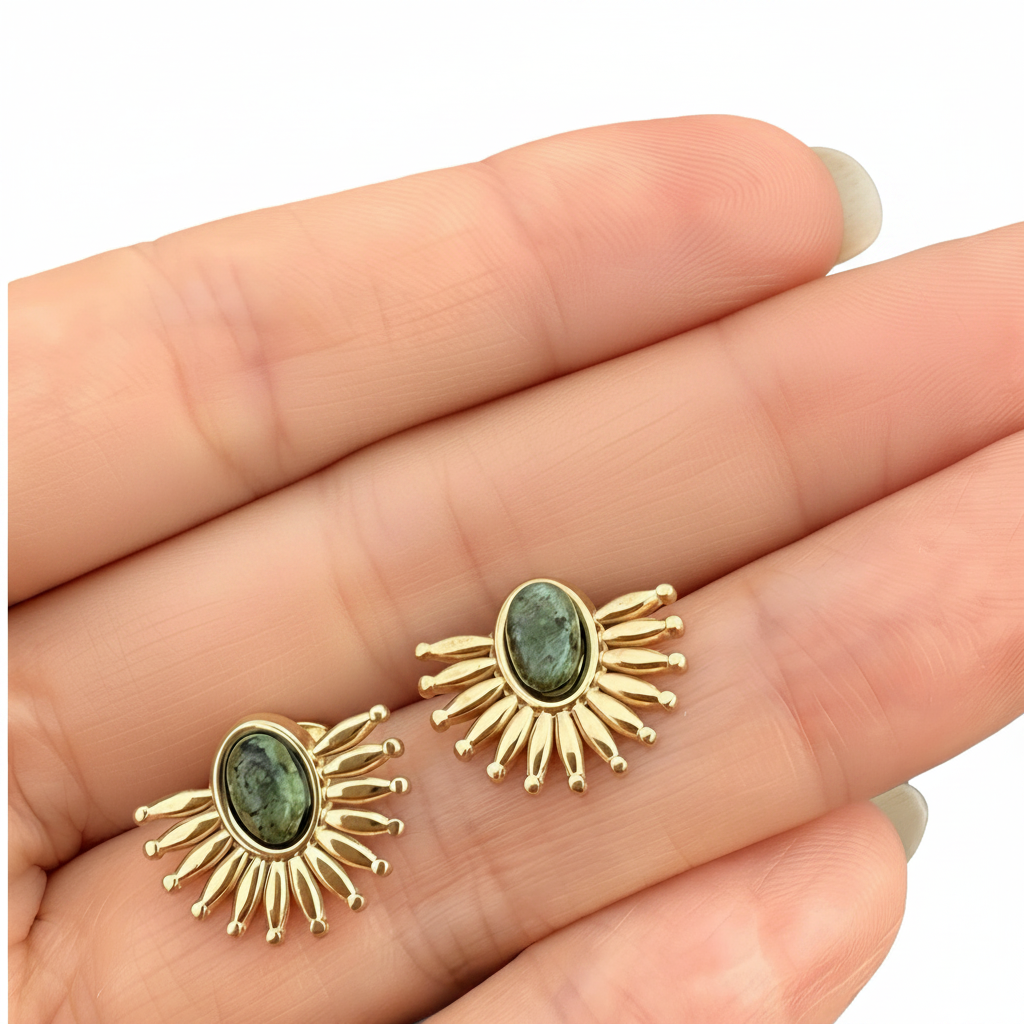 Gold sun-shaped earrings with green gemstones held between fingers on a white background