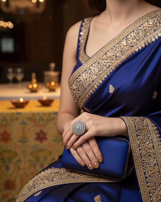 Woman in a blue saree with gold border holding a blue clutch in a decorated indoor setting.