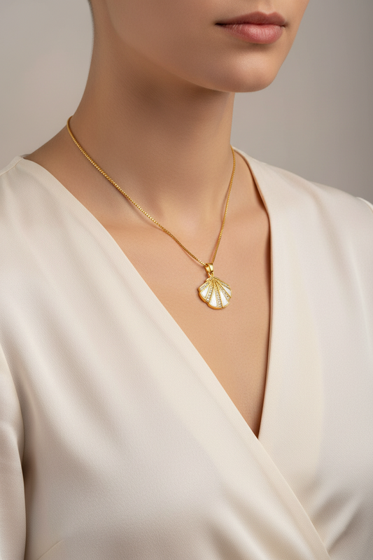 Gold necklace with a shell pendant worn by a person in a white dress against a neutral background