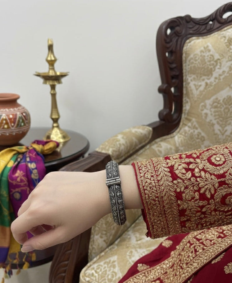 Hand wearing a bracelet with intricate design, sitting on a traditional chair with embroidered fabric.
