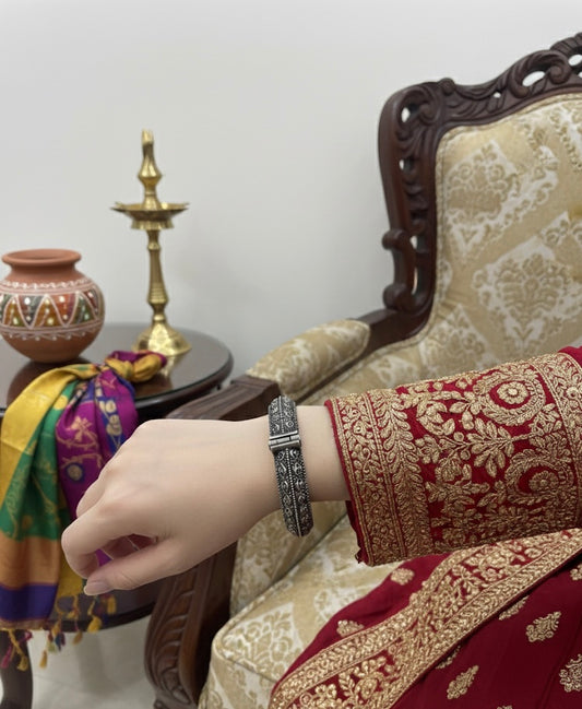 Hand wearing a bracelet with intricate design, sitting on a traditional chair with embroidered fabric.