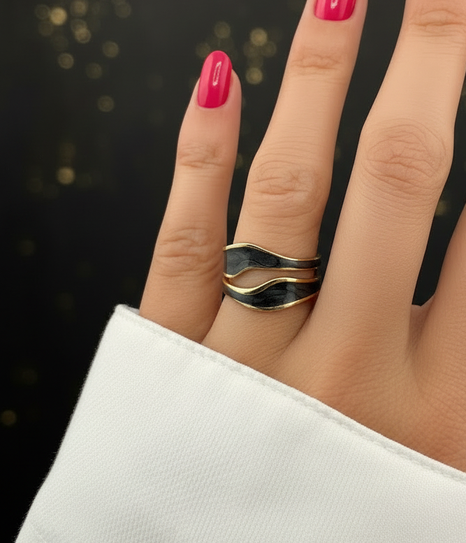 Hand wearing a gold and black ring with pink nail polish on a blurred background